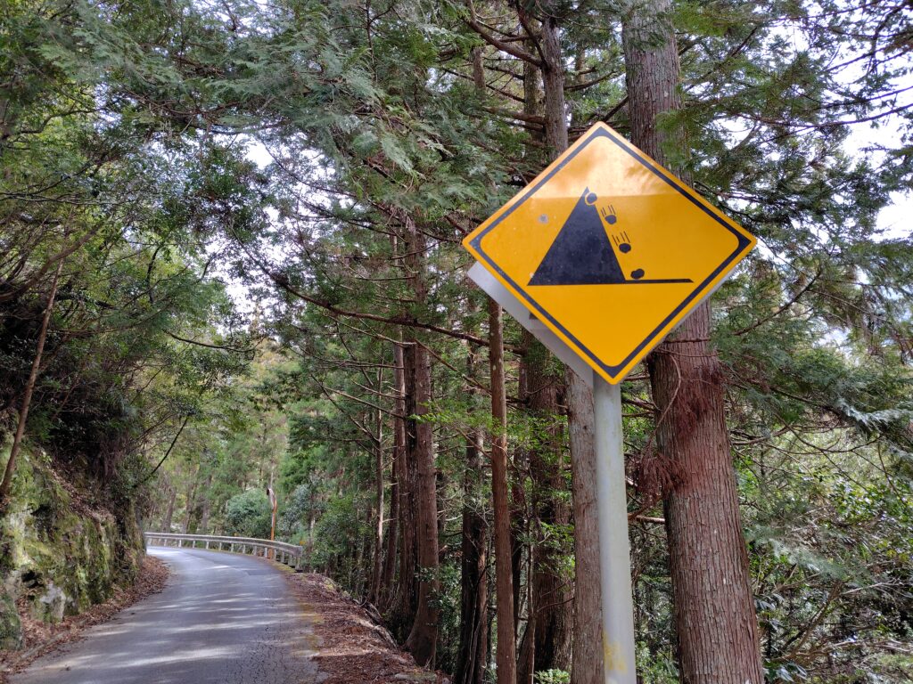 山林の狭い道路と落石注意標識がある場所不明土地のリスクを示すイメージ